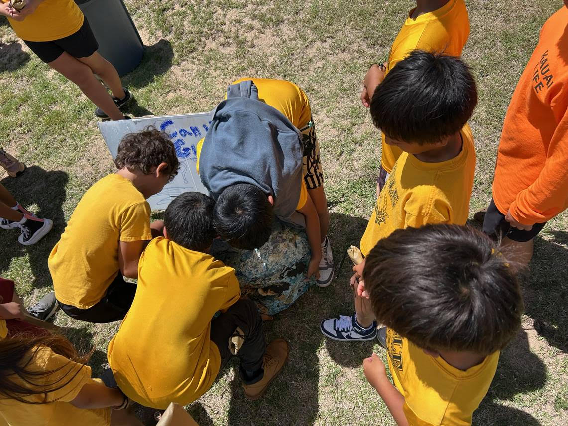 Students attempt to lift a ball of plastic waste collected from several debris clean-up efforts in order to guess its weight.